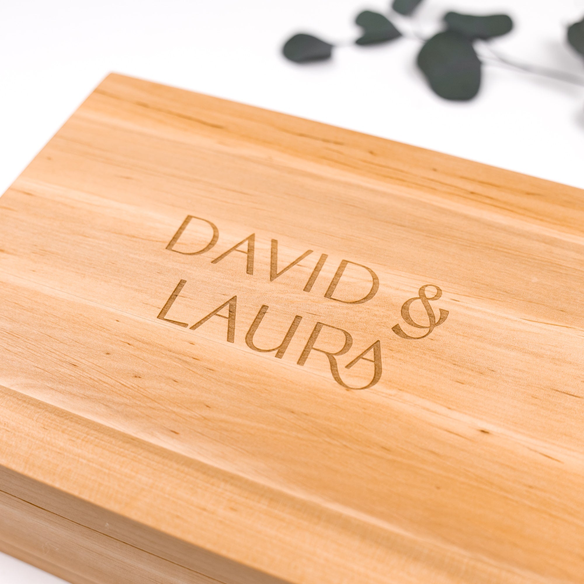 Wooden box with 'DAVID & LAURA' engraved on it, with a blurred green leaf in the background.
