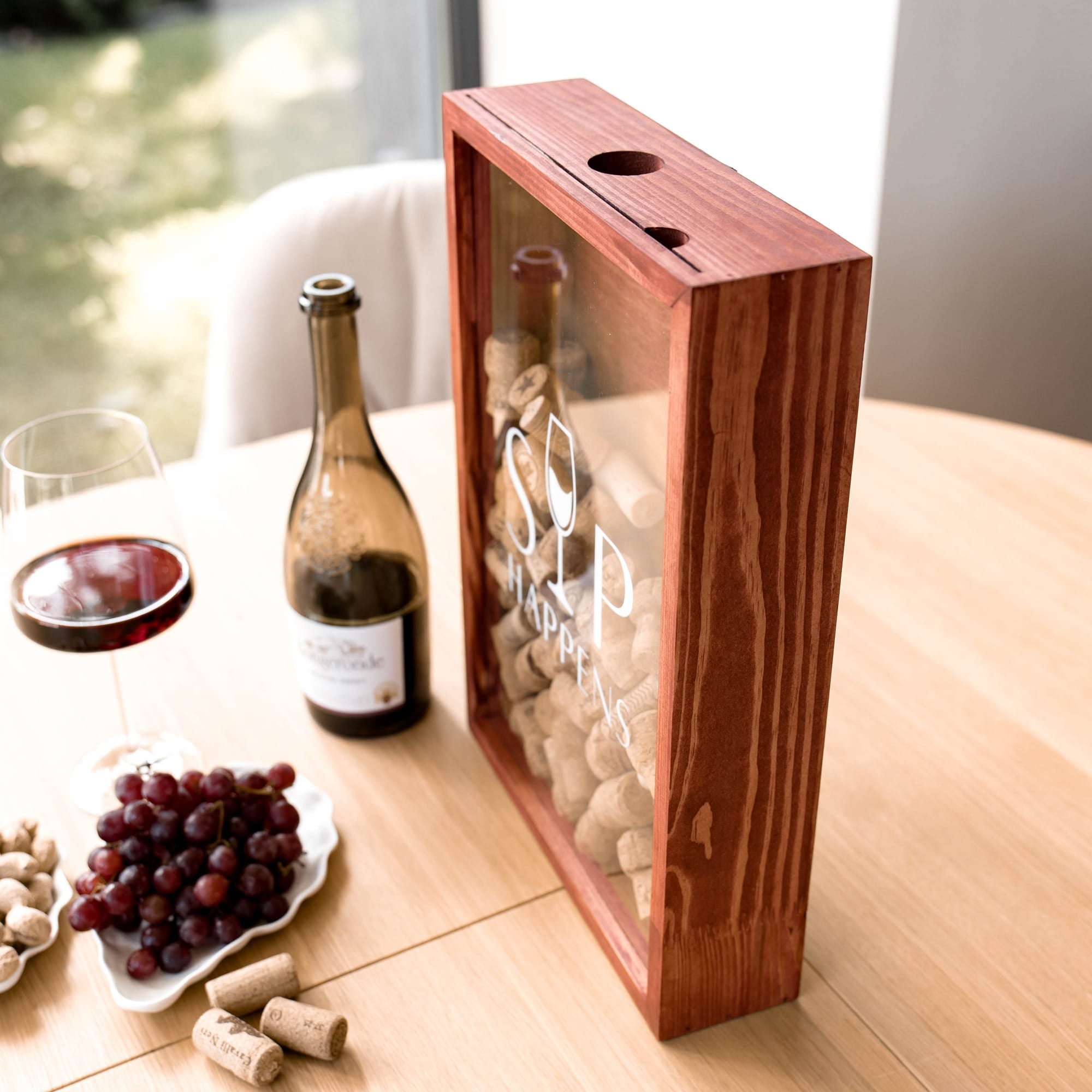 A wooden wine bottle storage box with clear front panels stands on a light wood table beside a bottle of sparkling wine, a glass of red wine, grapes, and corks. 