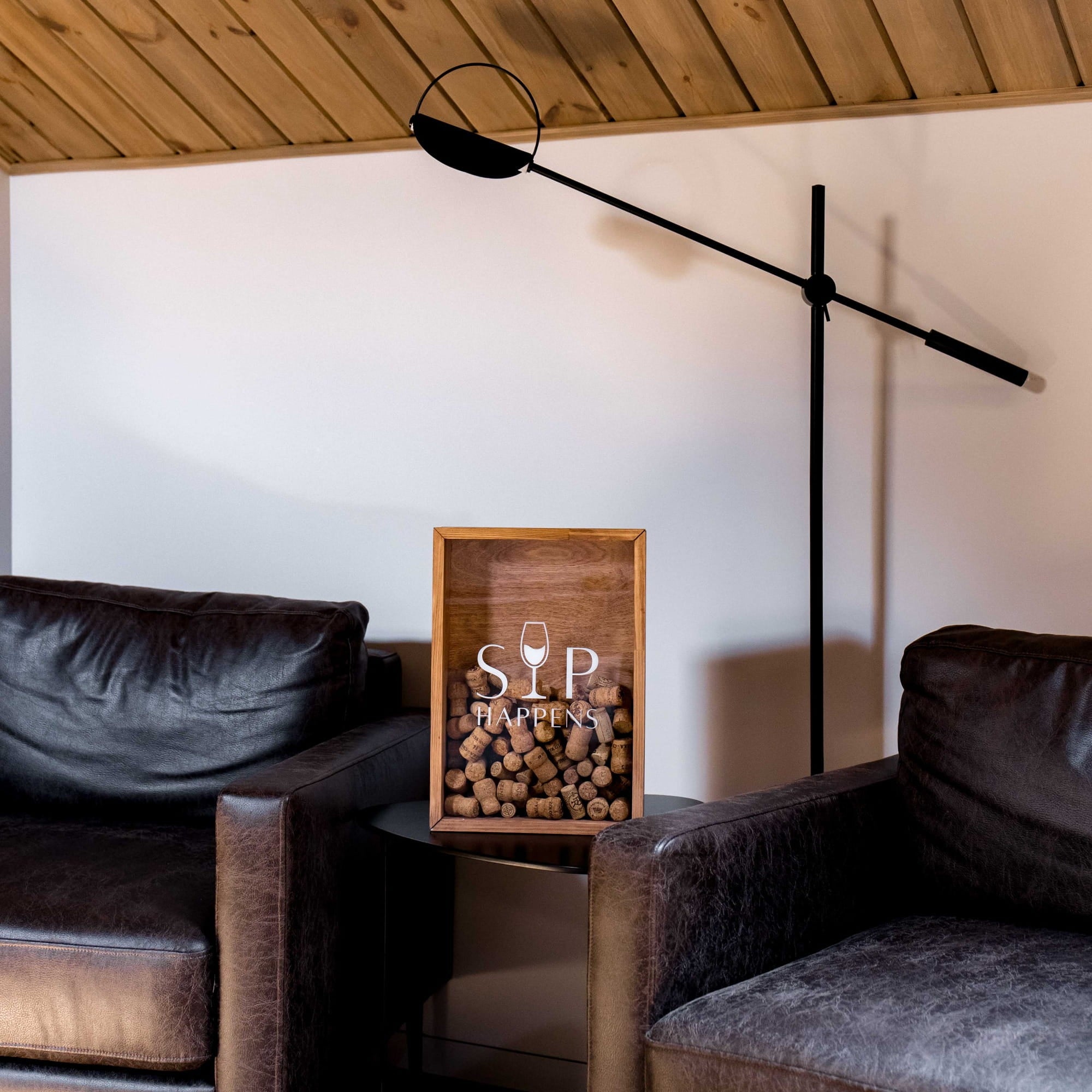 A framed cork barrel sign with 'SIP TUMBLERS' and a wine glass logo, styled on a side table between two dark leather armchairs in a modern living room setting.