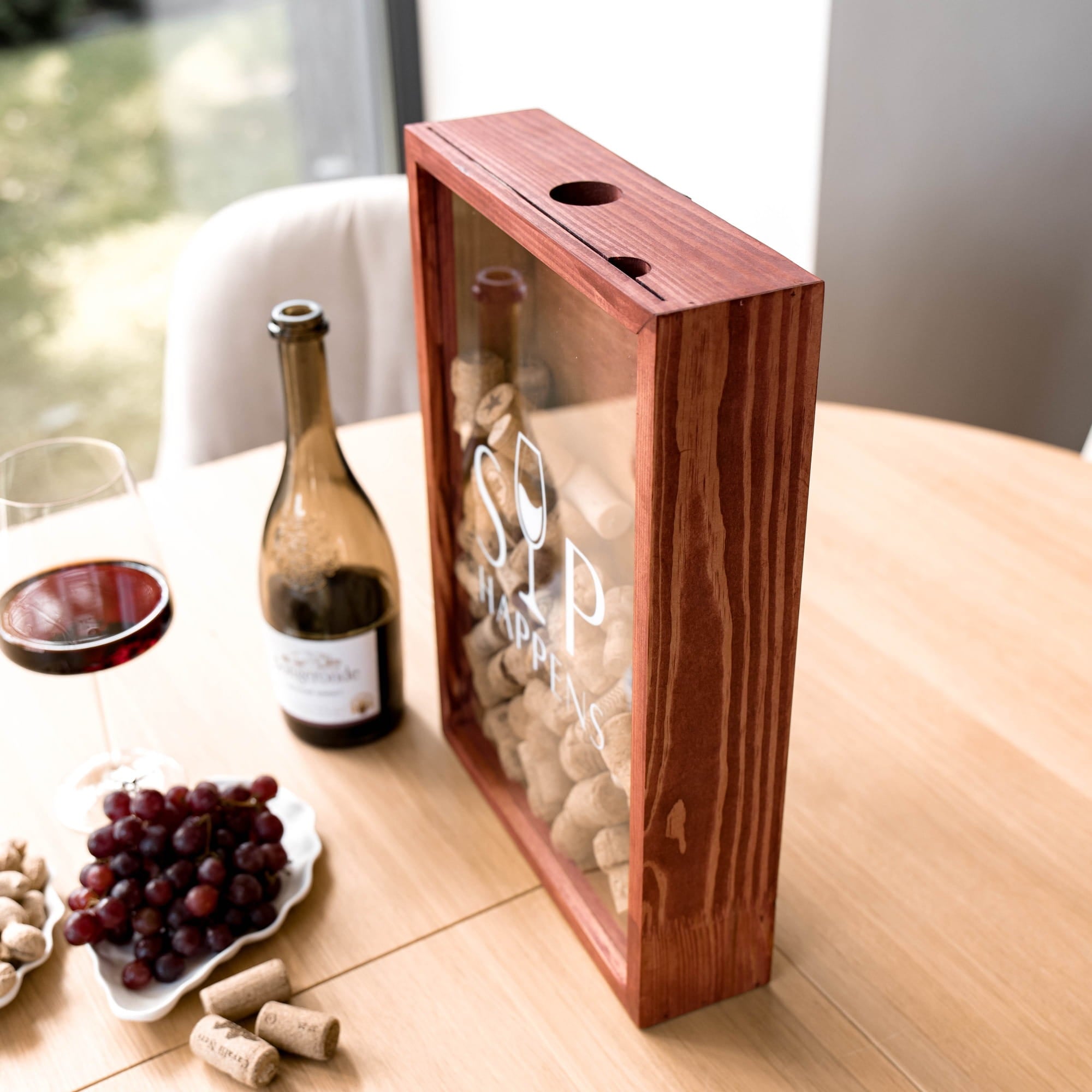 A wooden wine gift box with a clear front panel, shown on a light wood table beside a bottle of red wine, a glass of red wine, grapes, and corks. The box has a reddish-brown wood frame and a transparent front.