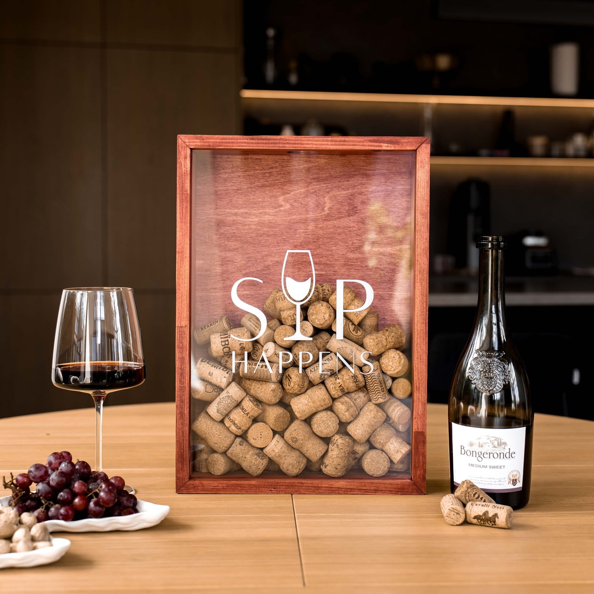 A framed wooden display filled with wine corks and featuring the text 'SIP HAPPENS' on a glass panel, styled on a wooden table with a glass of red wine, grapes, and a bottle of wine.