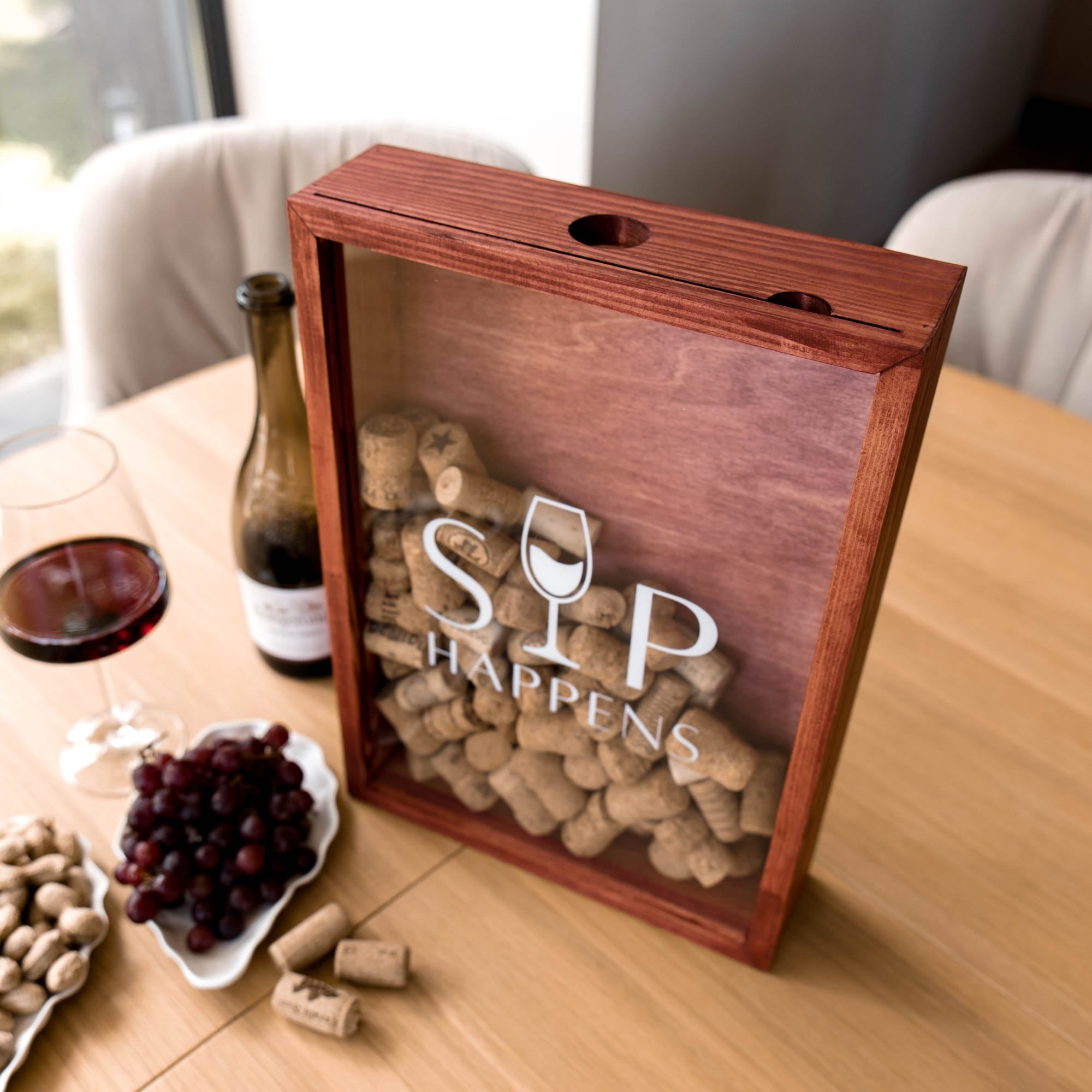 A wooden shadow box wine cellar with a clear front panel filled with wine corks and the words 'SIP HAPPENS' printed on it. The box sits on a wooden table next to a glass of red wine, a bottle of red wine, grapes, and pistachios.