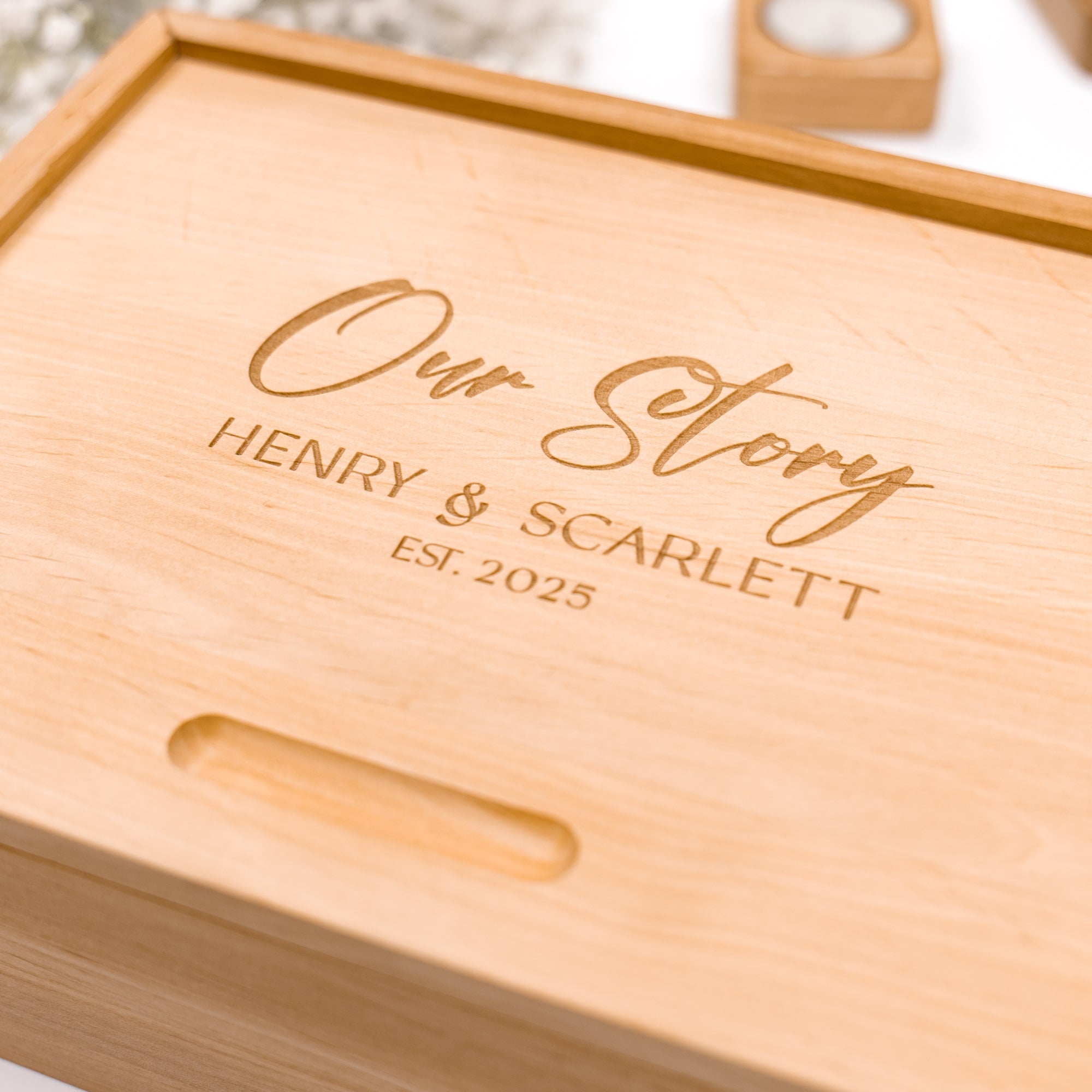 Wooden box with engraved text 'Our Story HENRY & SCARLETT EST. 2025' on a blurred background.