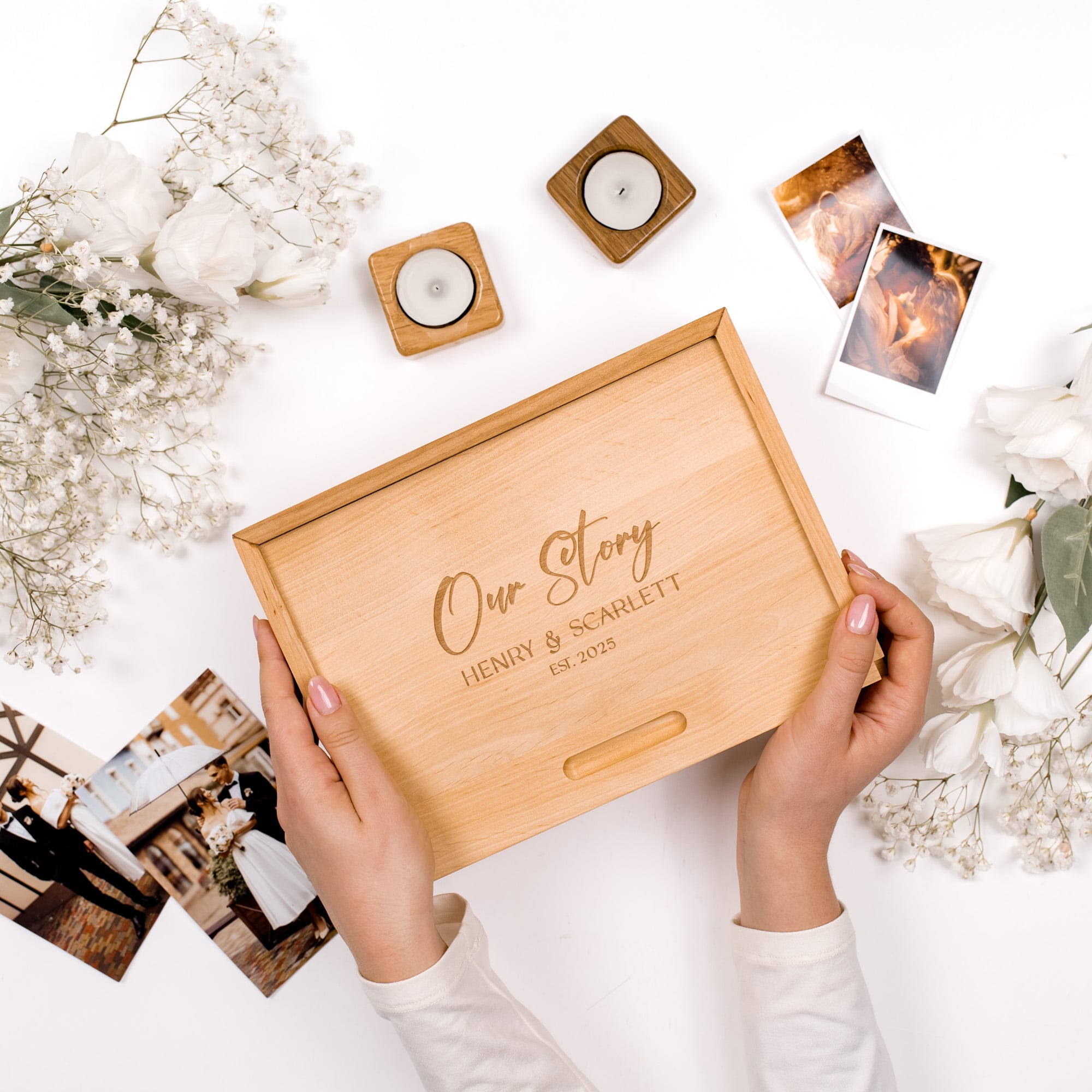 Wooden box with 'Our Story' engraving held by hands on a white surface with flowers and photos.