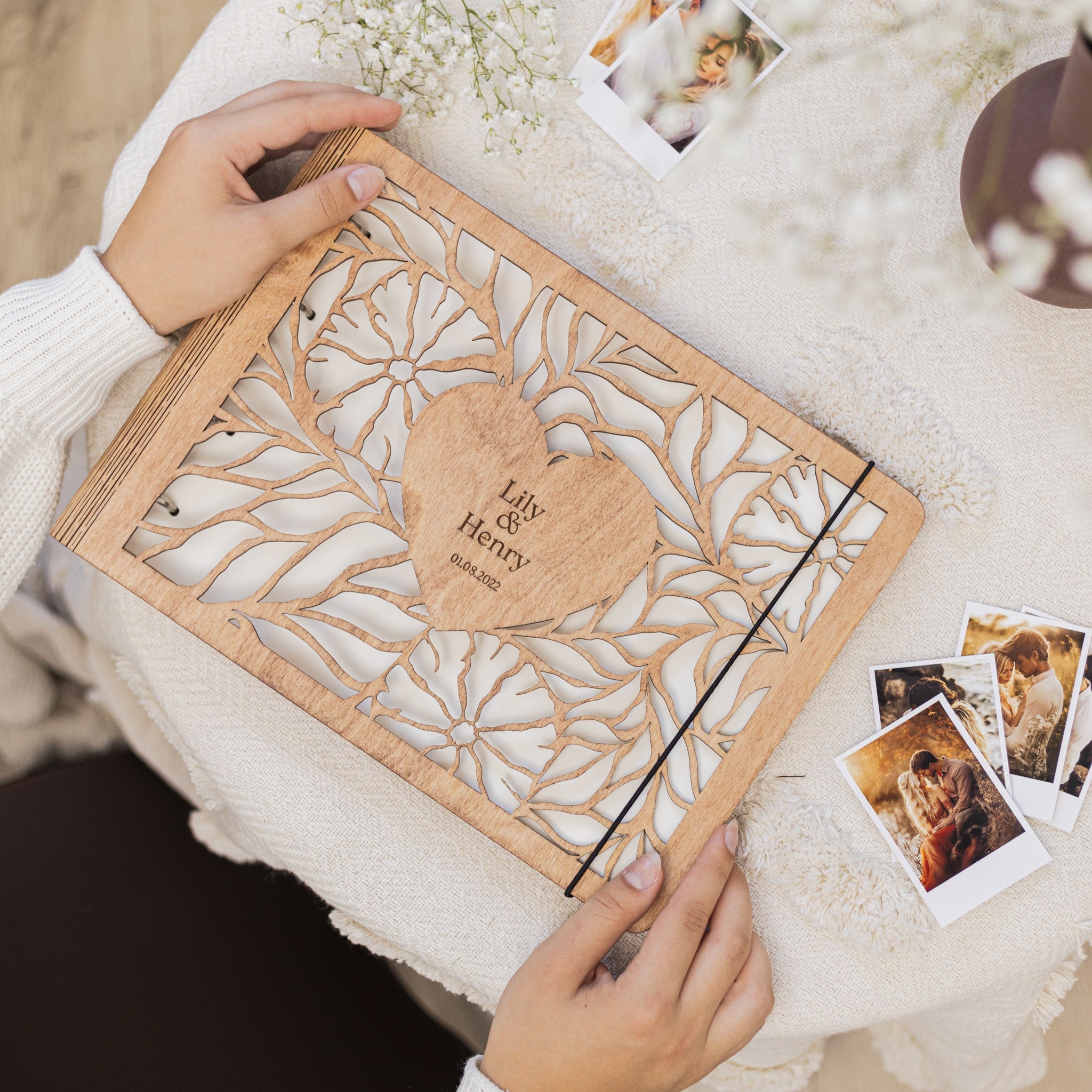 Wooden photo album with laser cut design and heart-shaped nameplate, held by a person on a soft surface with photos around.