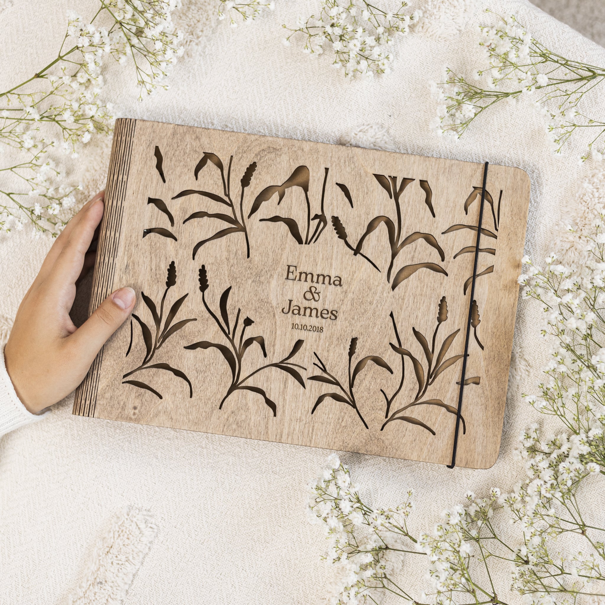 Wooden book with engraved floral design and text, held by a hand on a soft fabric background with flowers.