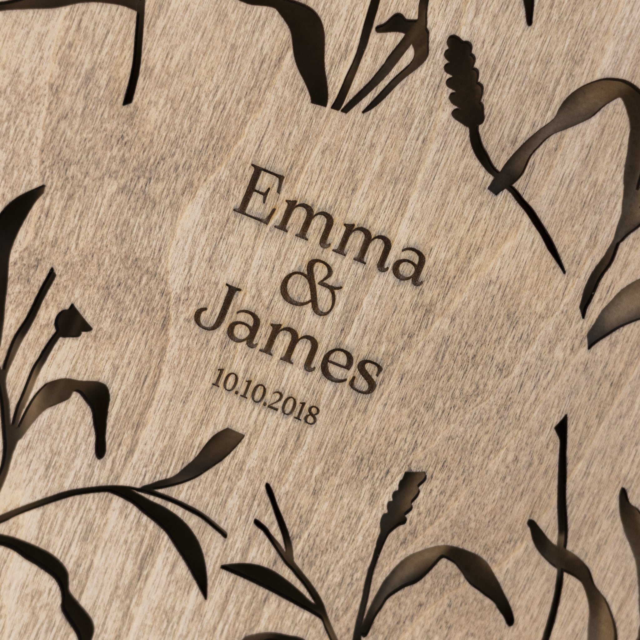 Wooden plaque with engraved names 'Emma & James' and date '10.10.2018', surrounded by black floral patterns.