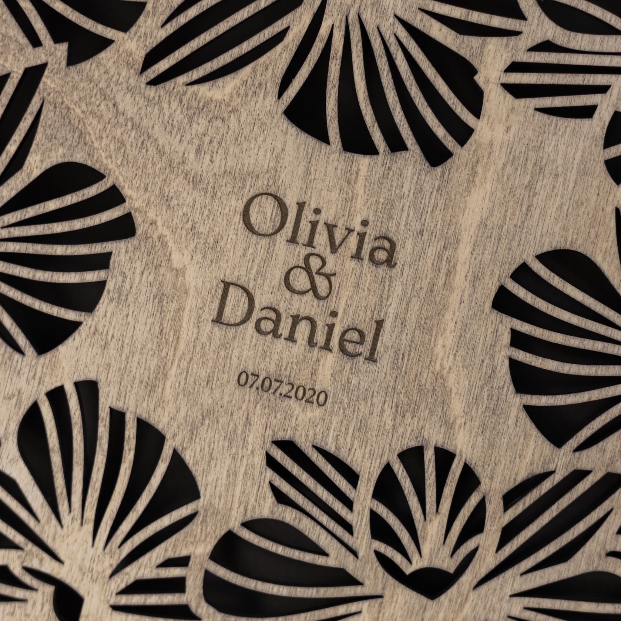 Wooden plaque with black leaf patterns and engraved text 'Olivia & Daniel 07.07.2020'.