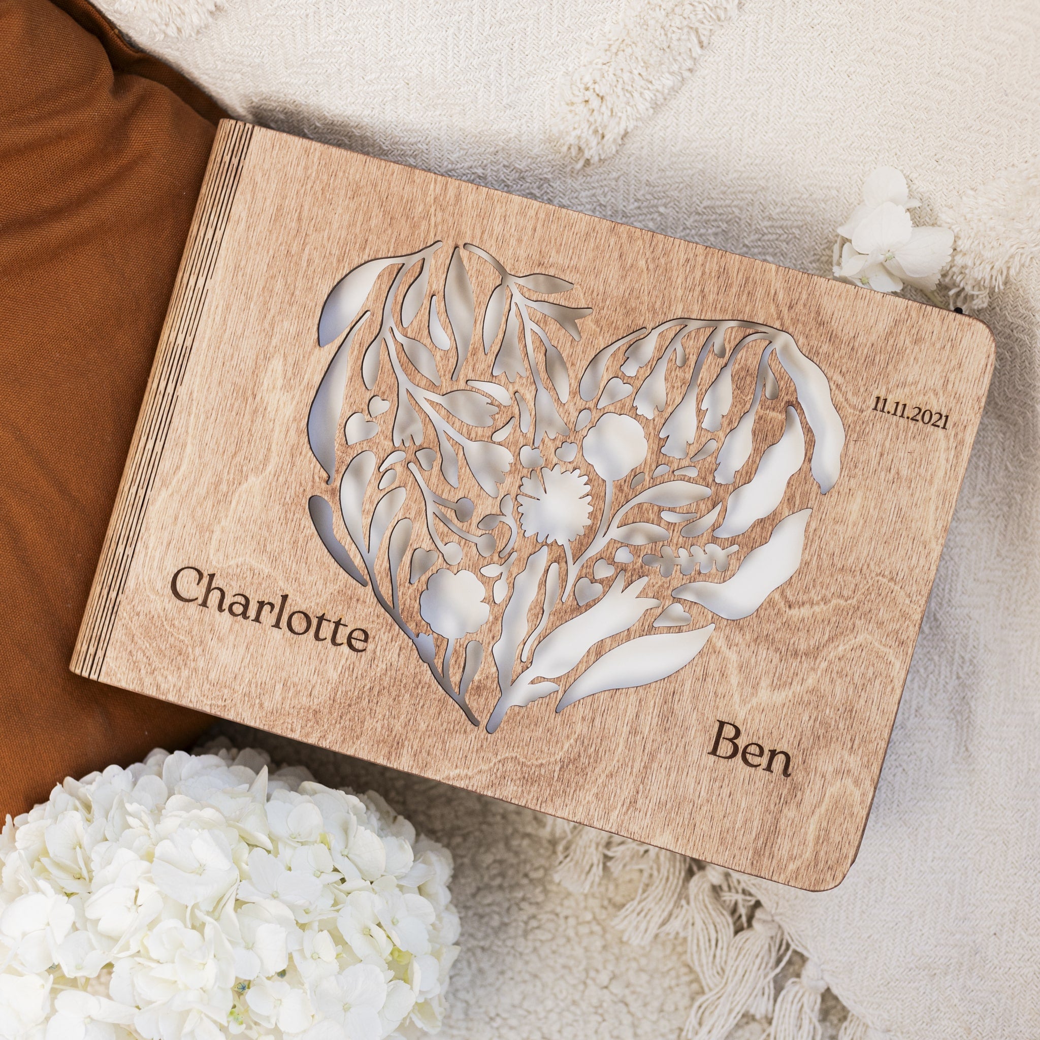 Wooden card with floral design and engraved names and date, surrounded by white flowers and brown fabric.