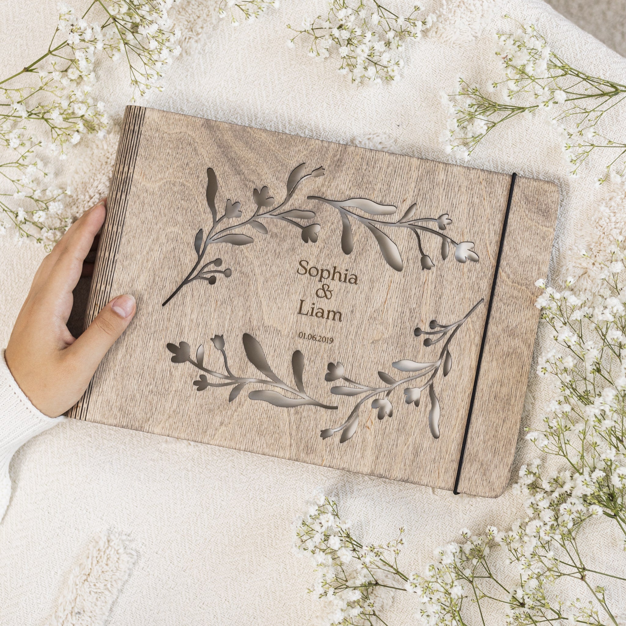 Wooden photo album with engraved names and date, surrounded by white flowers on a light background