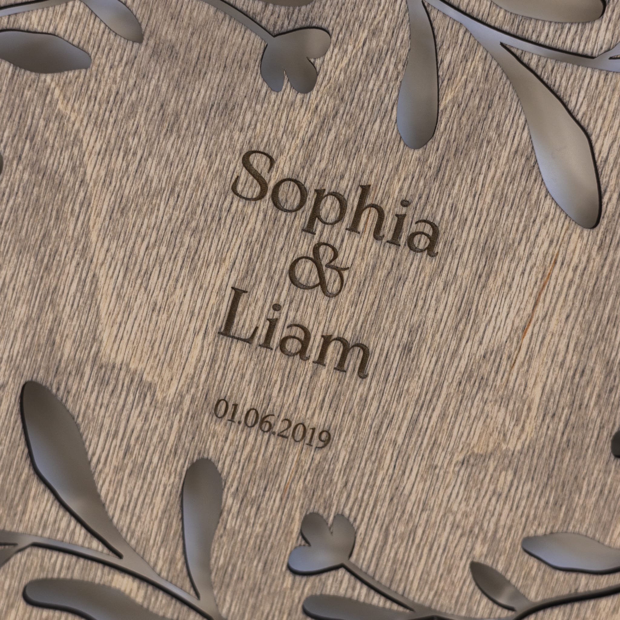 Wooden plaque with engraved names 'Sophia & Liam' and date '01.06.2019', surrounded by decorative leaves.