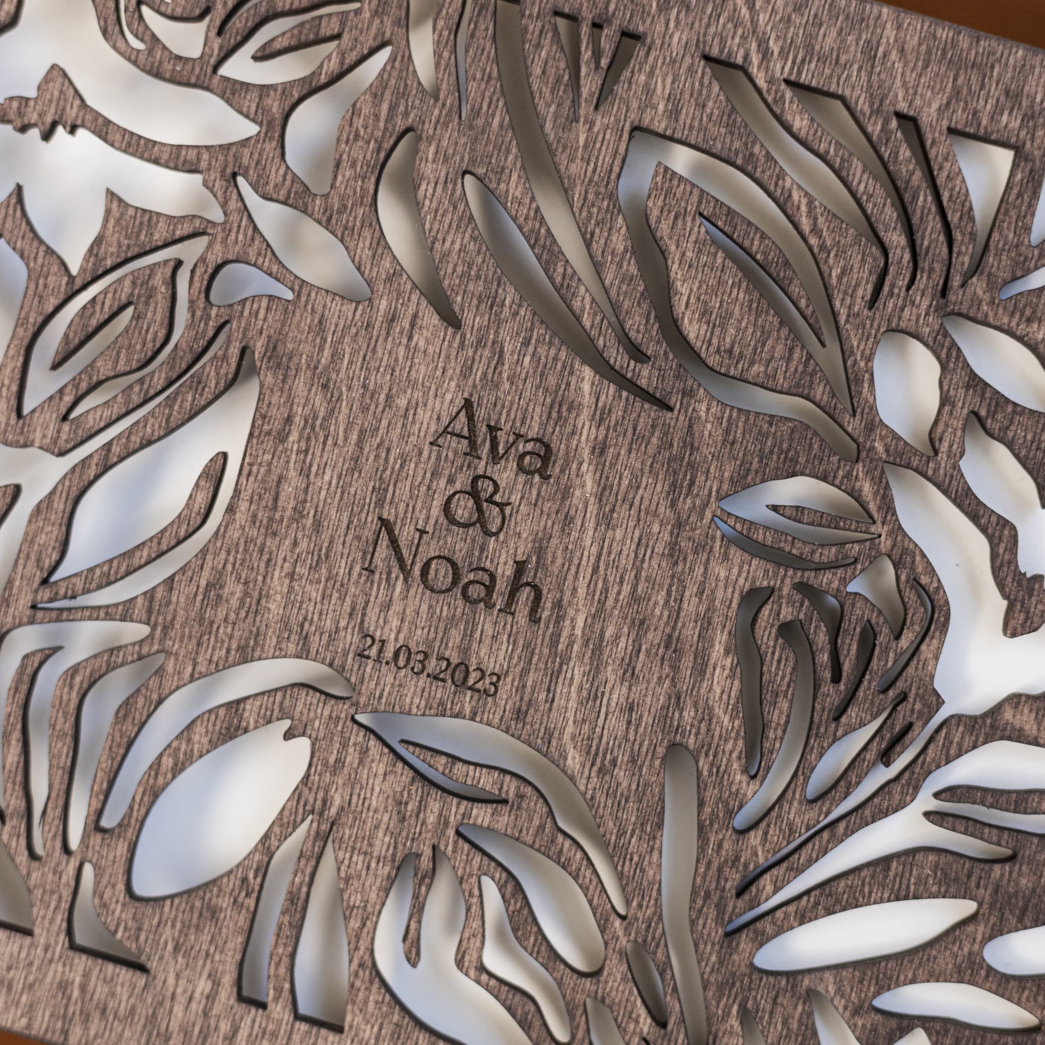 Wooden plaque with metal leaf designs and engraved text 'Ava & Noah 21.03.2023' on a wooden background.