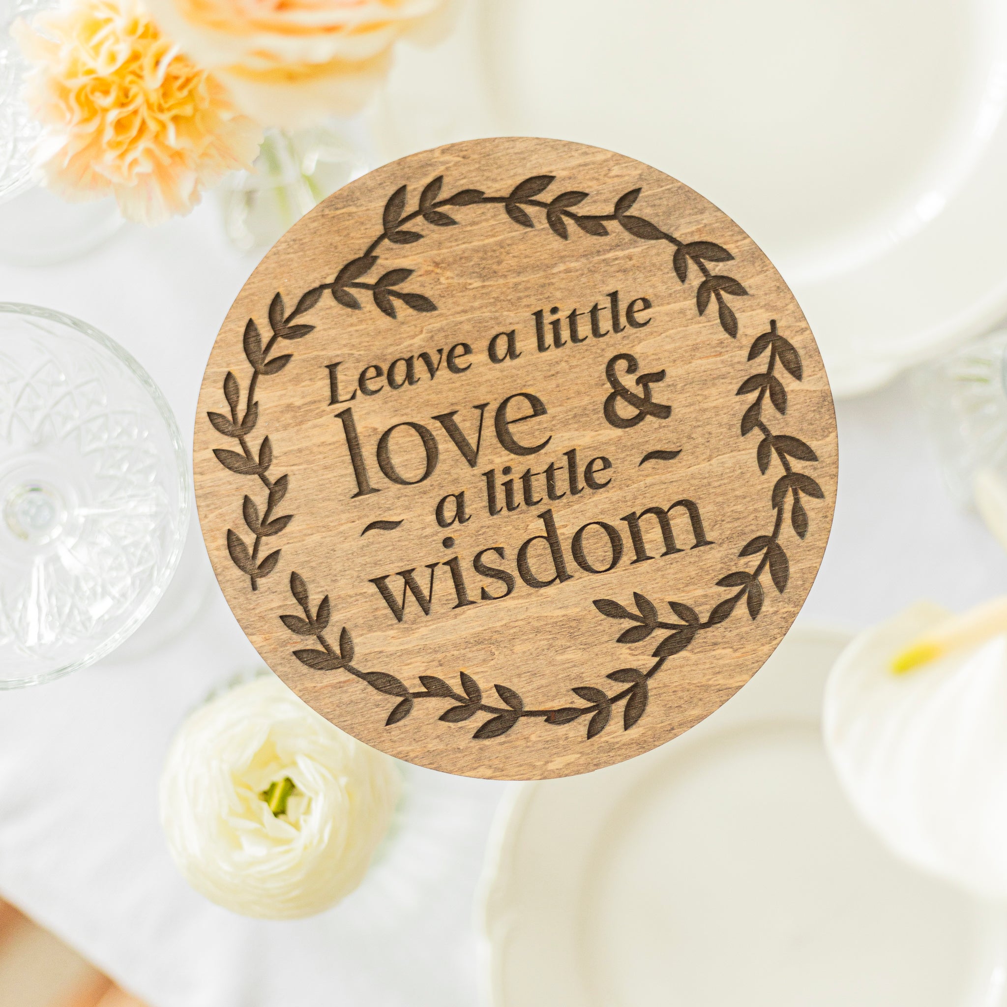 Wooden coaster with engraved text 'Leave a little love & a little wisdom' on a white surface with flowers.