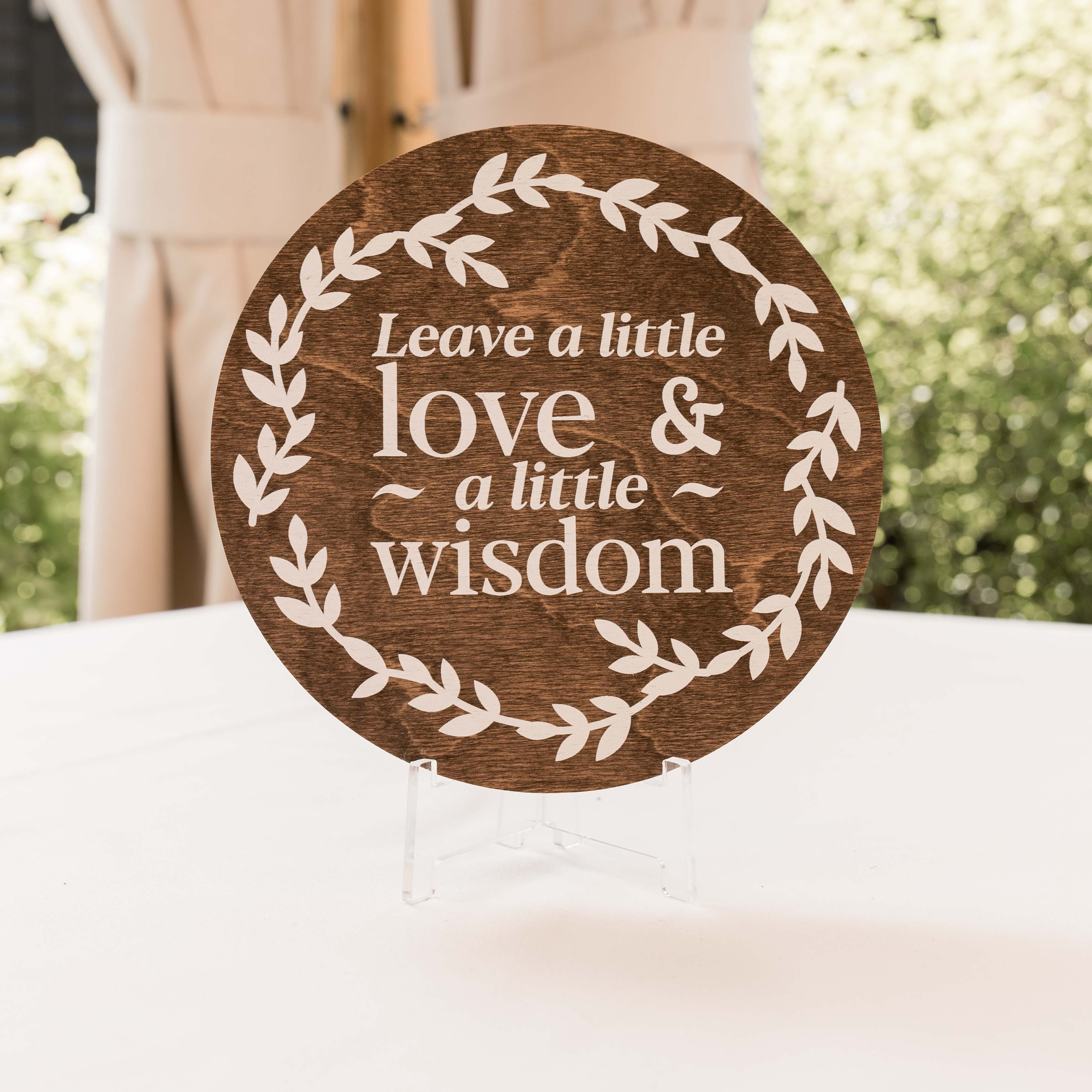Wooden plaque with 'Leave a little love & a little wisdom' text on a stand outdoors.
