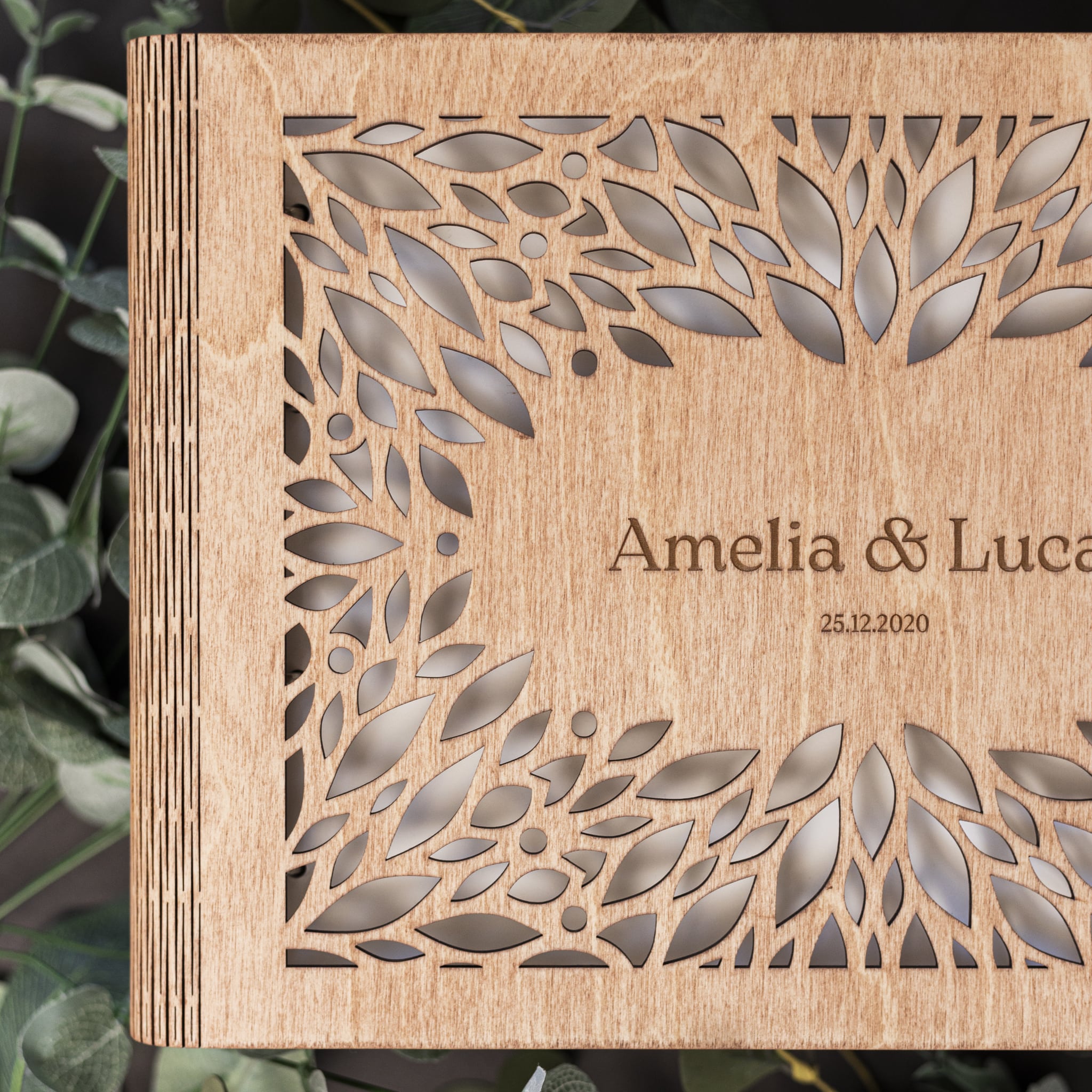 Wooden box with laser-cut leaf pattern and engraved text 'Amelia & Luca 25.12.2020' against a dark background.