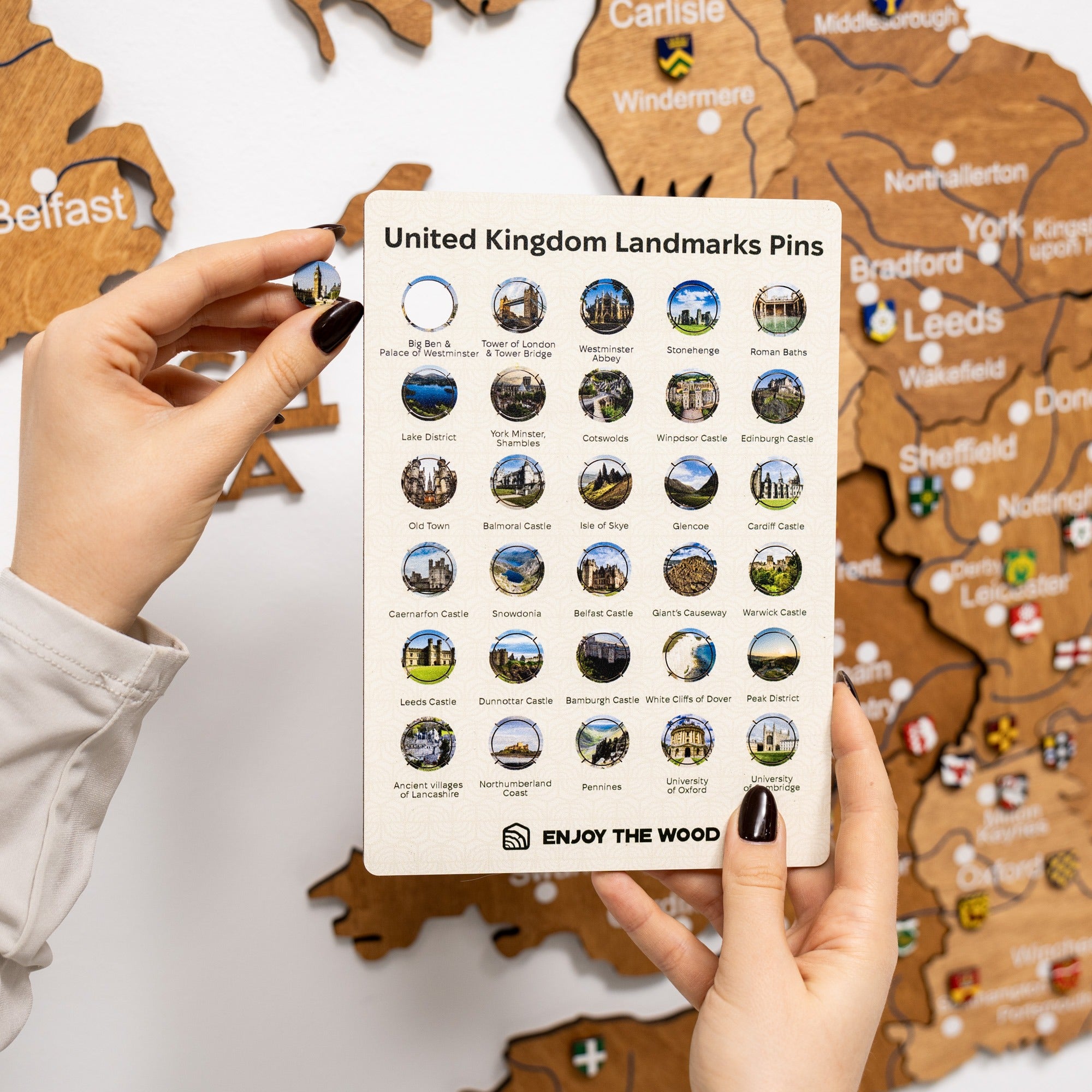 Hand holding a card with United Kingdom landmarks pins in front of a wooden map.