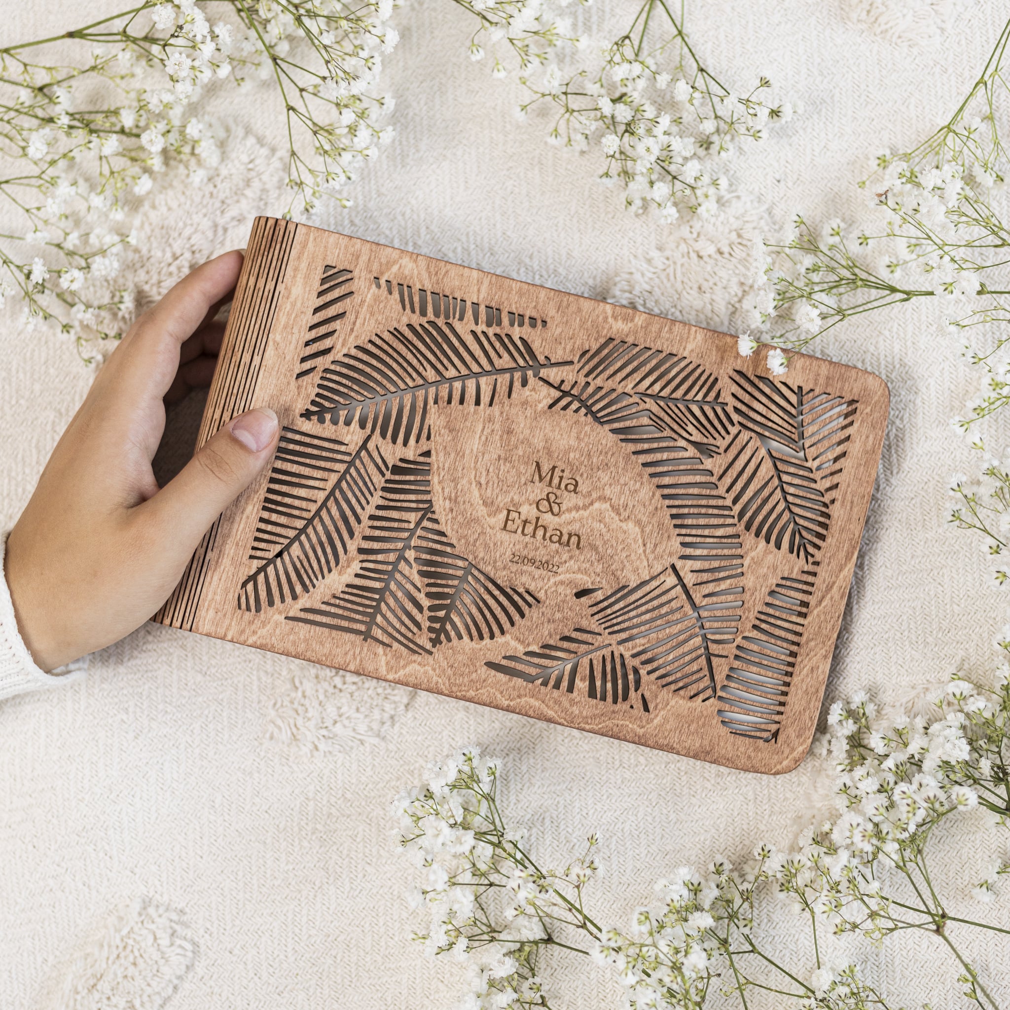 Wooden book with leaf patterns and engraved names, held by a hand on a light fabric background with white flowers.