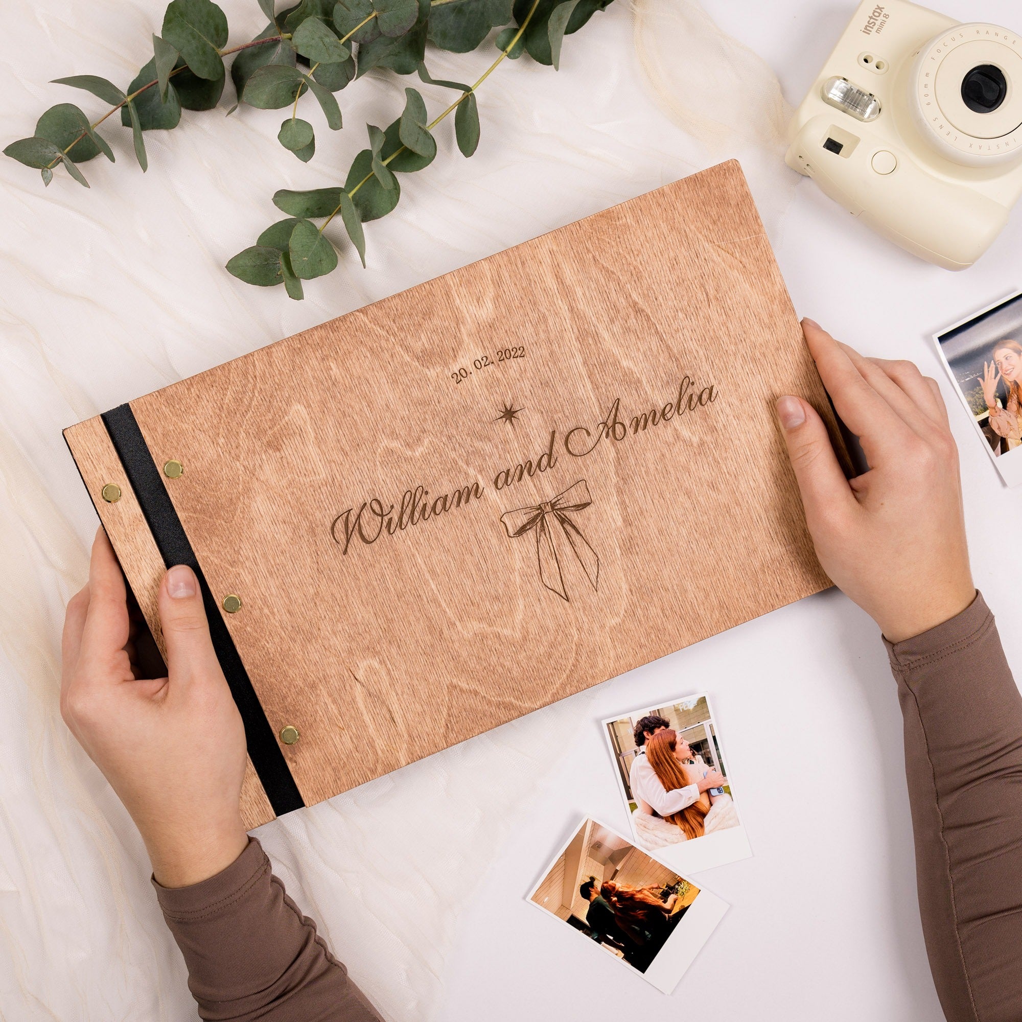 Personalized wooden photo album with engraved names, surrounded by photos and greenery on a white surface.