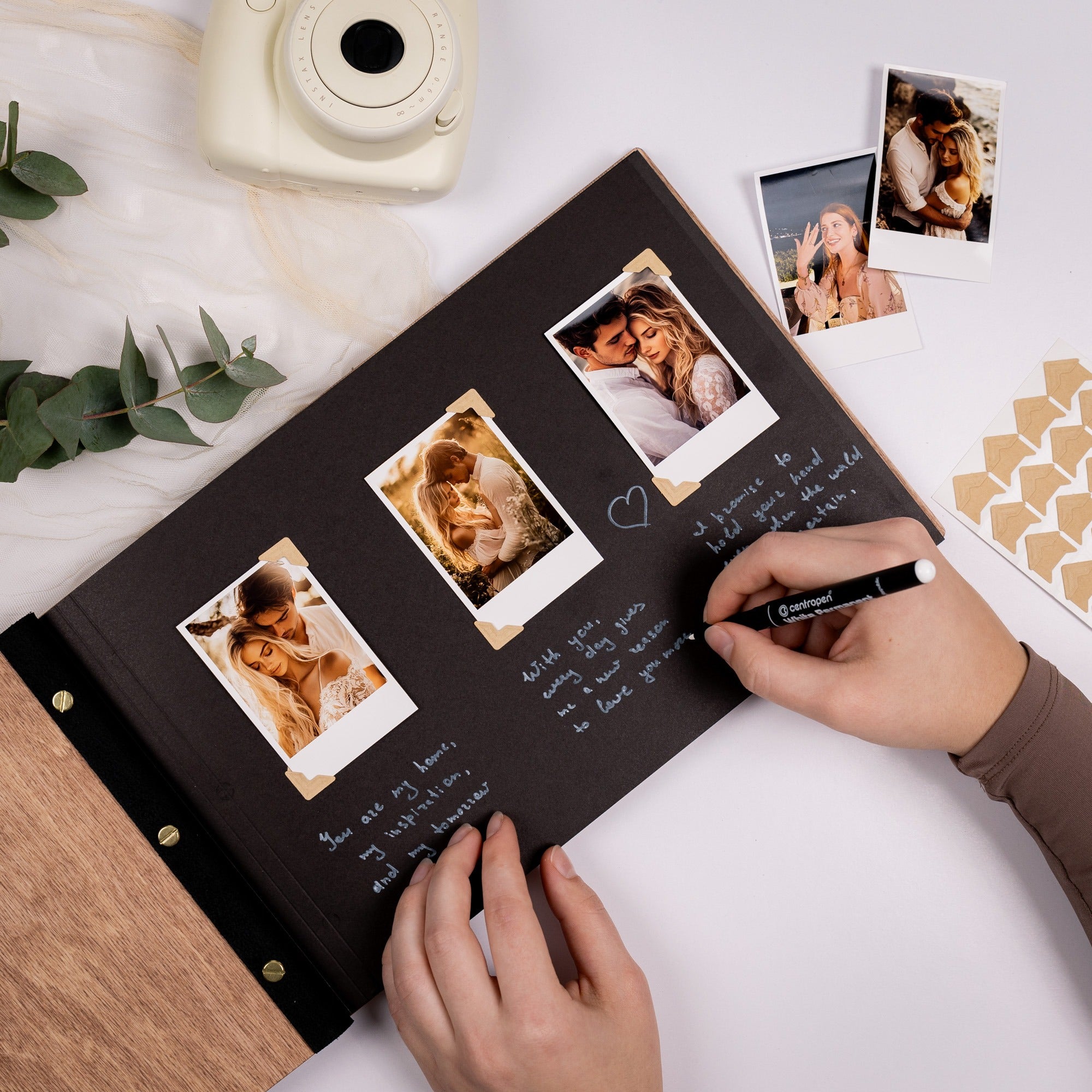 Person writing in a photo album with Polaroid-style prints and a camera on a white surface.