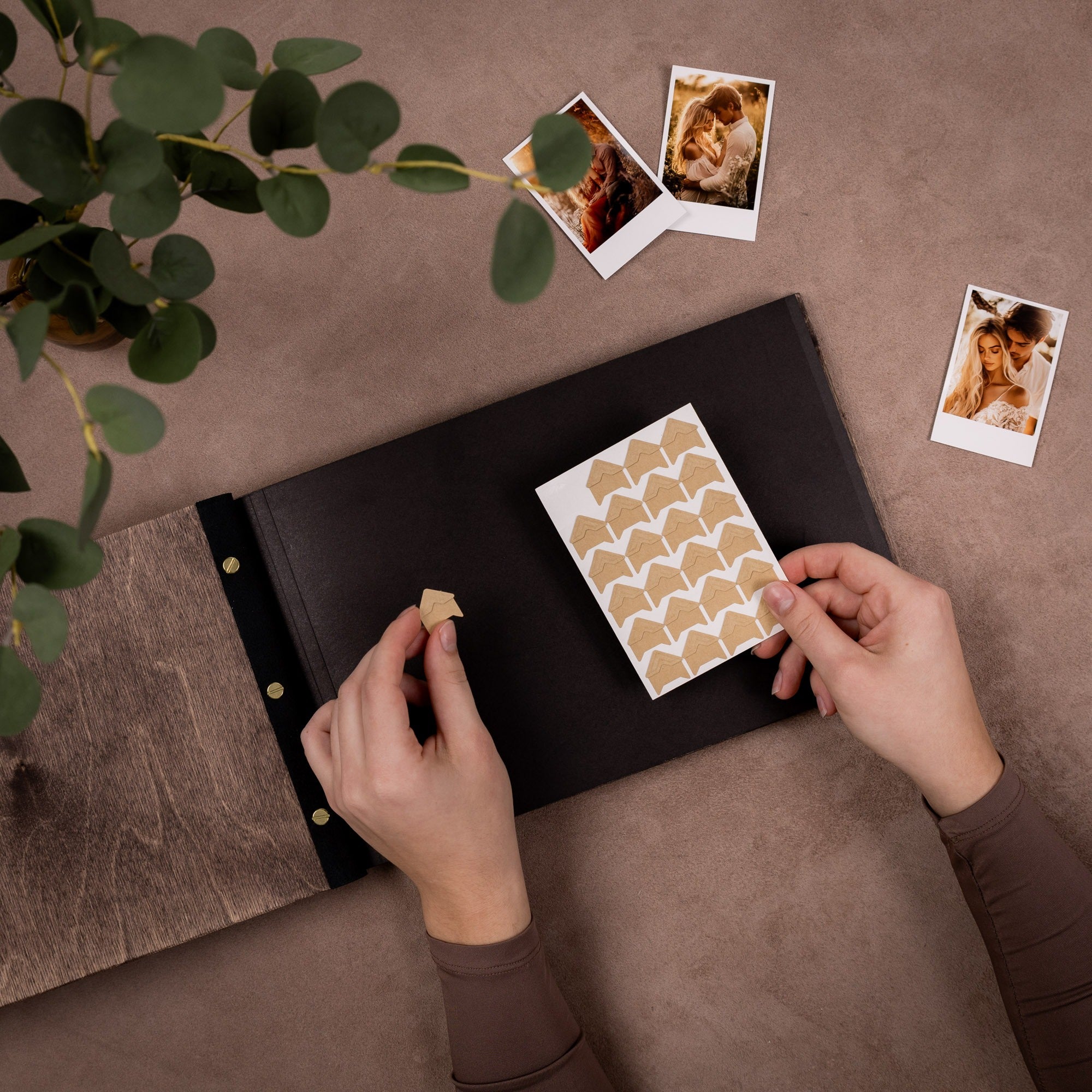 Person arranging heart-shaped stickers on a black photo album with scattered photos on a brown surface.