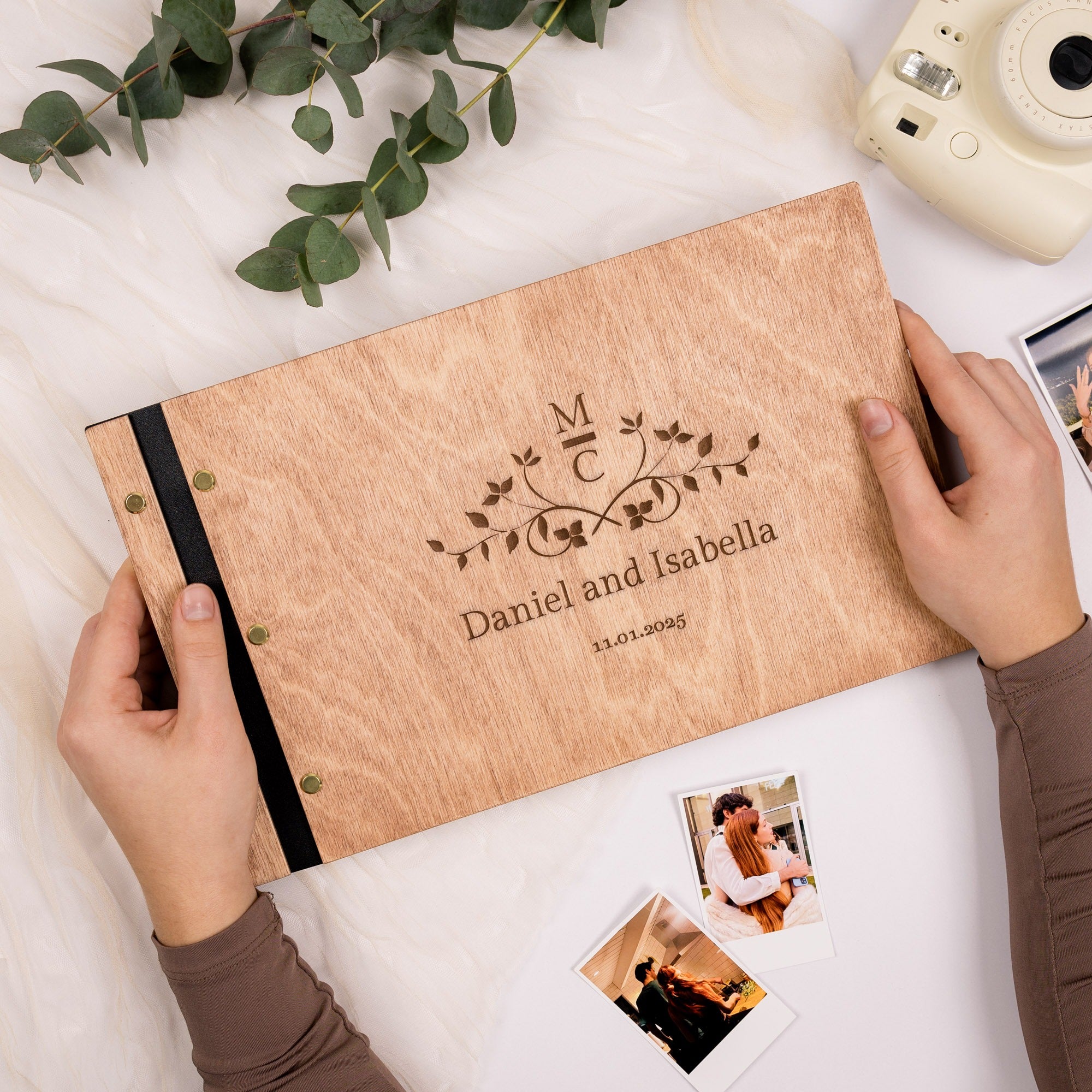 Wooden photo album with engraved names and dates, held by a person on a light surface with photos and greenery.