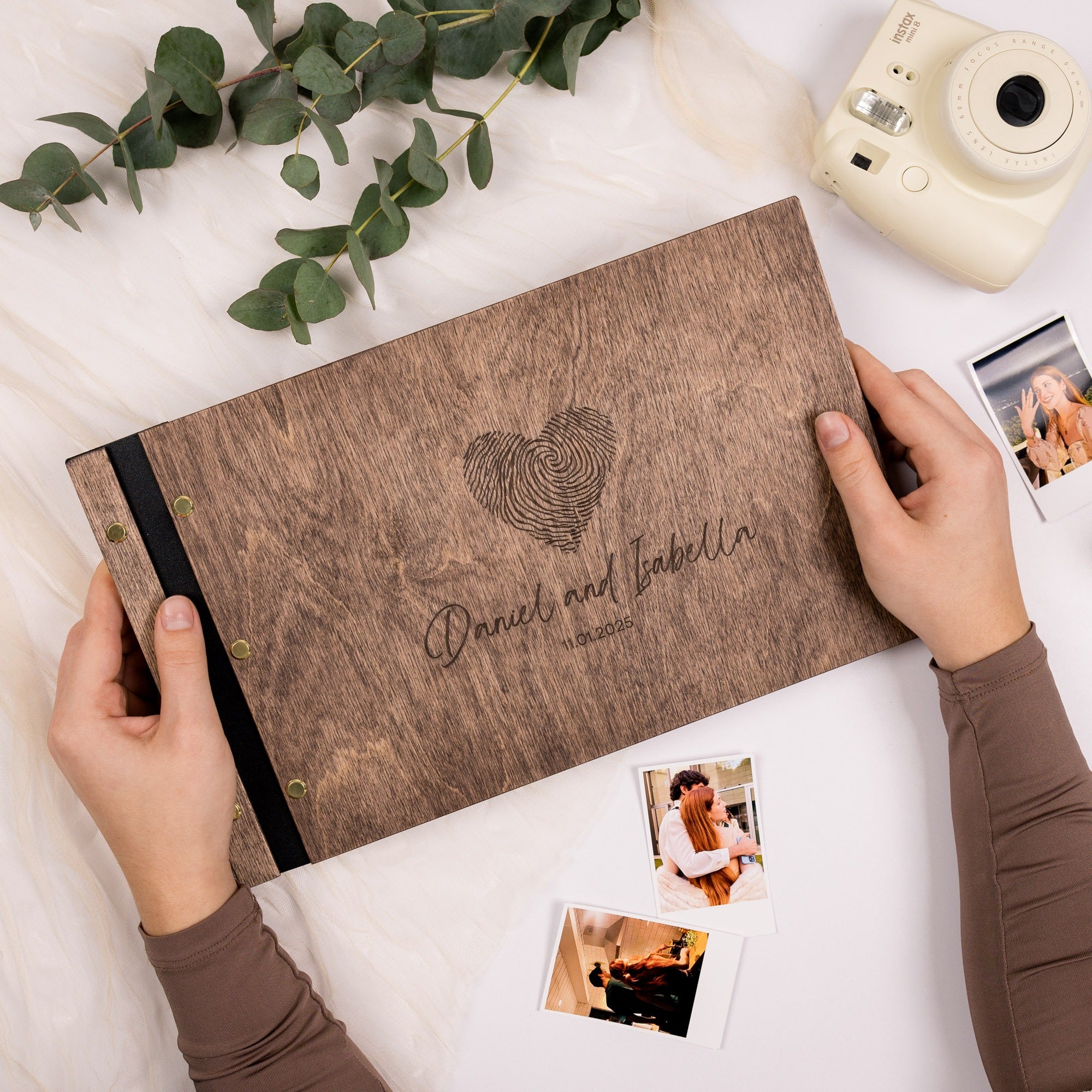 Wooden photo album with 'Daniel and Isabella' engraving, surrounded by photos and greenery on a white surface.