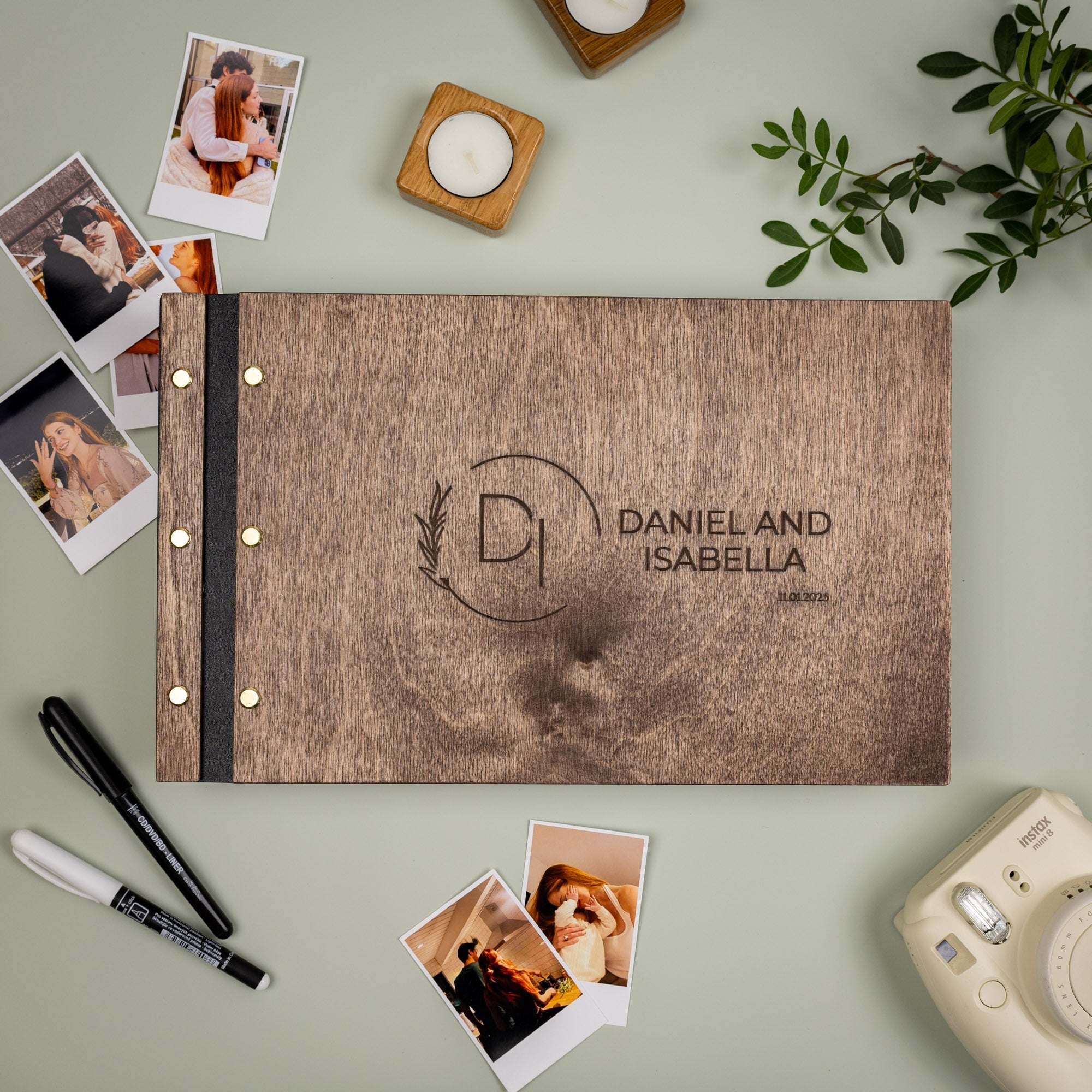 Wooden photo album with 'Daniel and Isabella' engraving, surrounded by Polaroid photos and a camera on a light green background.