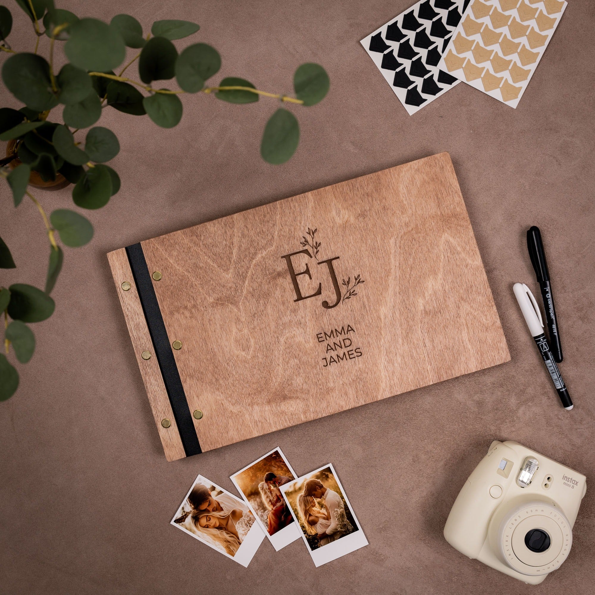 Personalized photo album with 'EJ' initials and 'Emma and James' text, surrounded by photos and a camera on a brown surface.