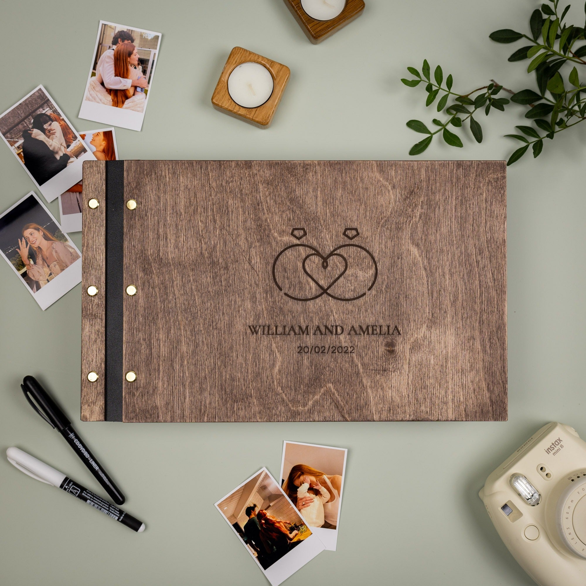 Wooden photo album with 'William and Amelia' engraving, surrounded by photos and a camera on a light green background.