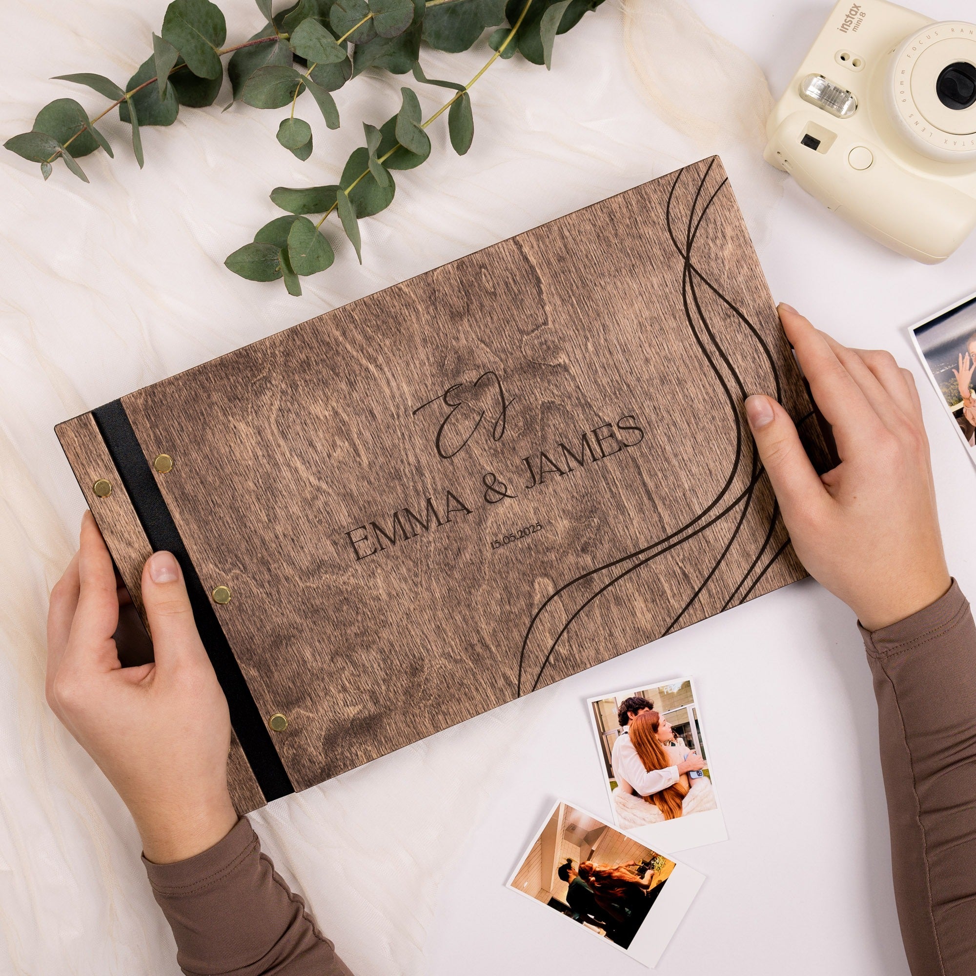 Wooden photo album with 'Emma & James' engraving, hands holding it, and photos on a light surface.