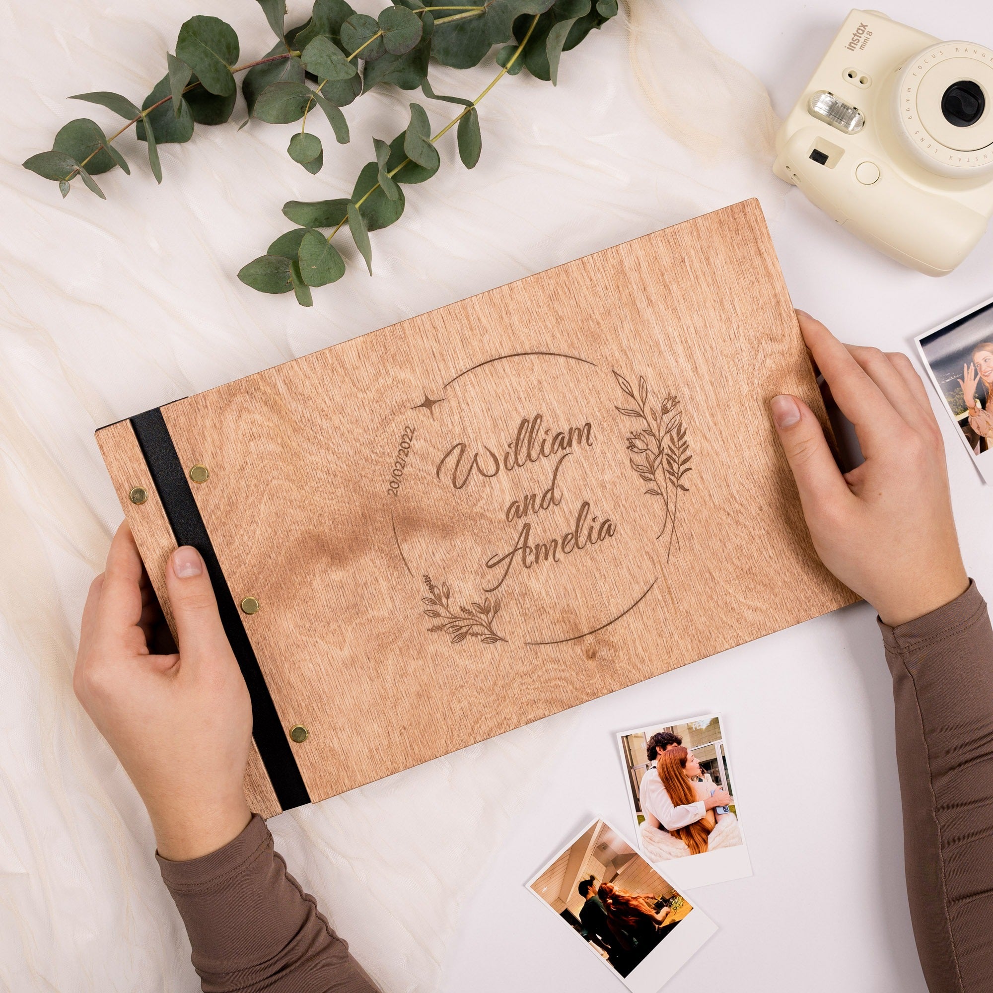 Personalized wooden photo album with engraved names on a white surface with greenery and a camera.
